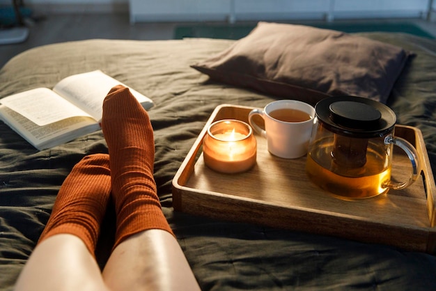 Foto suave de piernas de mujer con calcetines de lana en la cama con un libro, una taza de té y una vela en la bandeja