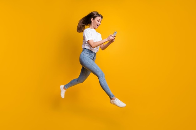 Foto de tamaño completo del cuerpo de la mujer influyente saltando navegando por internet con teléfono celular aislado sobre fondo de color amarillo brillante