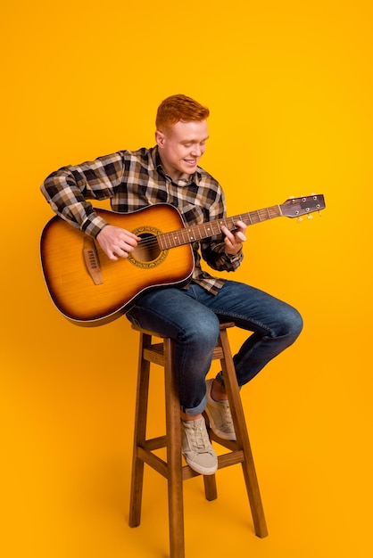 Foto a tamaño completo de un joven apuesto que se sienta en un taburete y toca la guitarra en un escenario aislado sobre un fondo de color amarillo