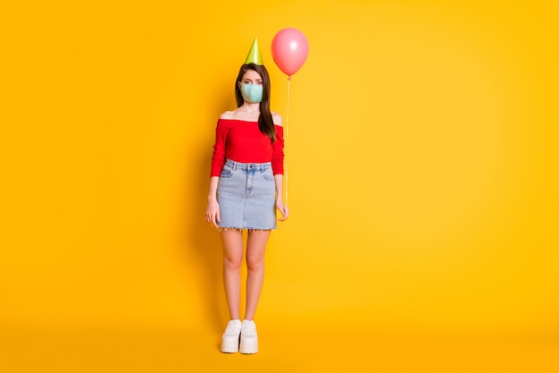 Foto de tamaño completo de niña con máscara médica tendencia de distancia social celebrar cumpleaños solo sostener globo usar falda de mezclilla superior roja piernas zapatillas de deporte aisladas sobre fondo de color brillante