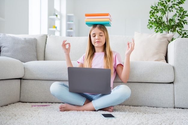 Foto de tamaño completo de un niño pequeño enfocado, estudio remoto, computadora portátil, sostenga la pila, libros, materiales de la lección, cabeza, medite antes de la comunicación en línea, siéntese con las piernas cruzadas y dobladas en casa en el interior