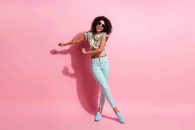 Foto de tamaño de cuerpo entero de una mujer de piel negra feliz descuidada bailando en el piso con gafas de sol de traje casual aislado sobre fondo de color rosa pastel