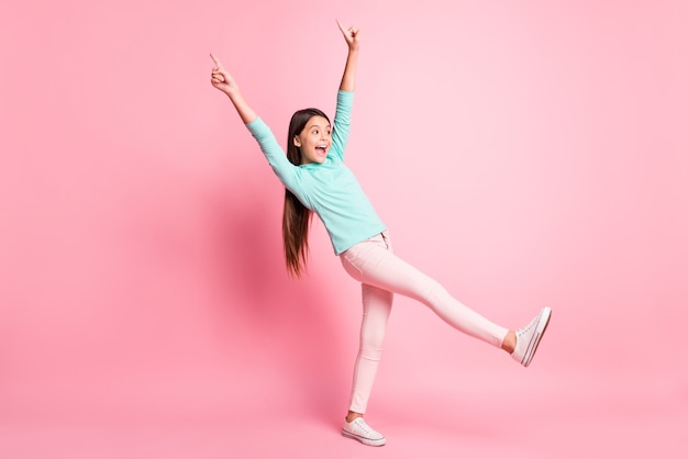 Foto del tamaño del cuerpo de longitud completa de una pequeña niña juguetona apuntando hacia arriba con los dedos avanzando sonriendo aislado sobre fondo de color rosa