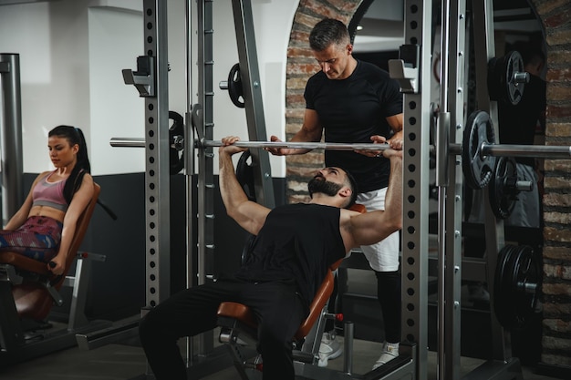 Una foto de un tipo musculoso con ropa deportiva haciendo ejercicio con un entrenador personal en el gimnasio. Está bombeando músculos del pecho durante un entrenamiento duro.