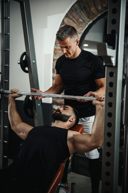 Una foto de un tipo musculoso con ropa deportiva haciendo ejercicio con un entrenador personal en el gimnasio. Está bombeando músculos del pecho durante un entrenamiento duro.