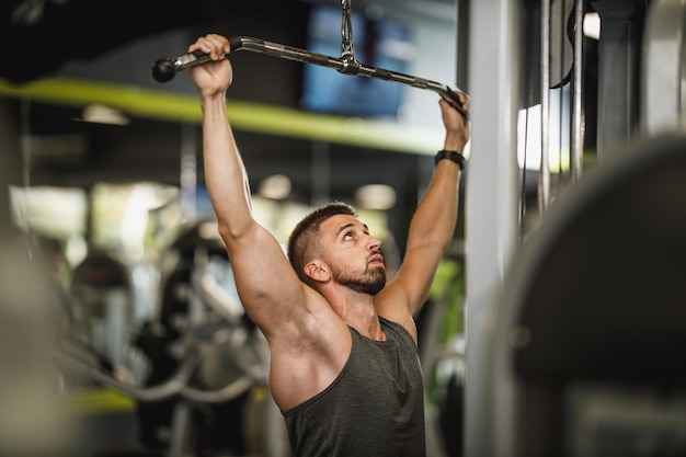 Una foto de un tipo musculoso con ropa deportiva haciendo ejercicio en la máquina del gimnasio. Está bombeando los músculos de la espalda con mucho peso.