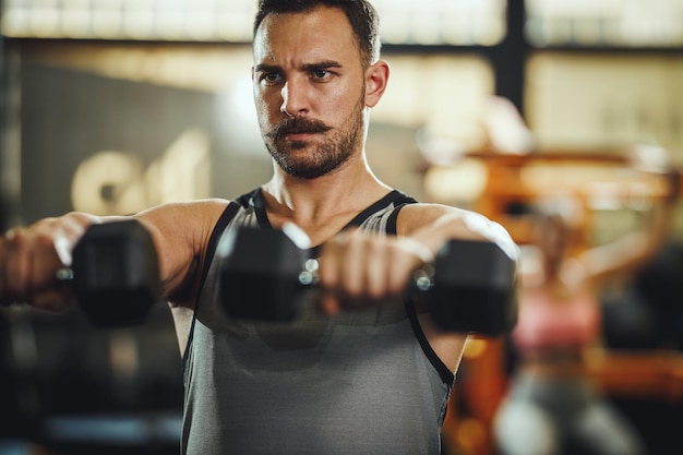 Una foto de un tipo musculoso con ropa deportiva haciendo ejercicio con pesas en el gimnasio de entrenamiento cruzado. Está bombeando los músculos de los hombros con mucho peso.