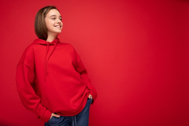 Foto tiro de atraente feliz positivo sorridente morena adolescente feminina vestindo elegante capuz vermelho em pé isolado sobre a parede de fundo vermelho, olhando para o lado. espaço vazio, copie o espaço