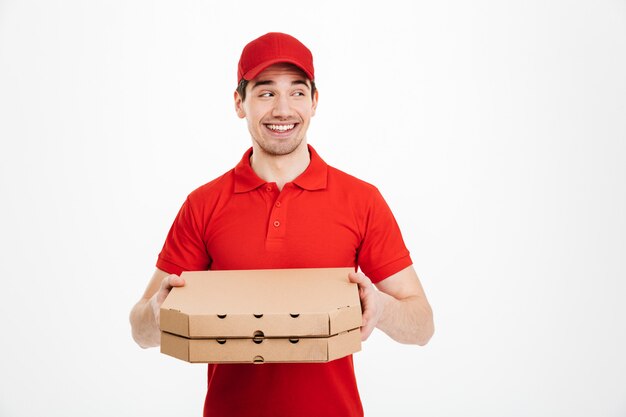 Foto del trabajador del servicio de entrega en camiseta roja y gorra sosteniendo dos cajas de pizza para llevar y mirando a un lado en copyspace, aislado sobre un espacio en blanco