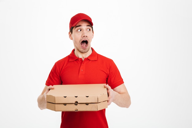 Foto del trabajador del servicio de entrega en camiseta roja y gorra sosteniendo dos cajas de pizza para llevar y mirando a un lado en copyspace con sorpresa, aislado sobre un espacio en blanco