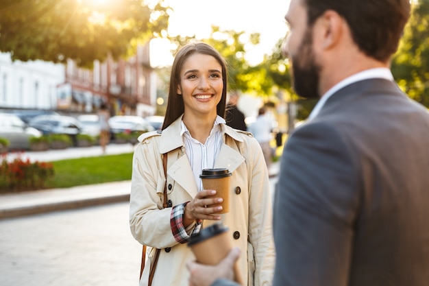 Foto de trabajadores de oficina complacidos hombre y mujer en ropa formal bebiendo café para llevar y mirándose mientras hablan en las calles de la ciudad