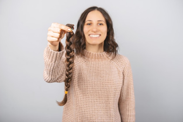 Foto de una trenza de cabello moreno para pacientes con cáncer sostenida por una niña sonriente en un estudio sobre fondo gris