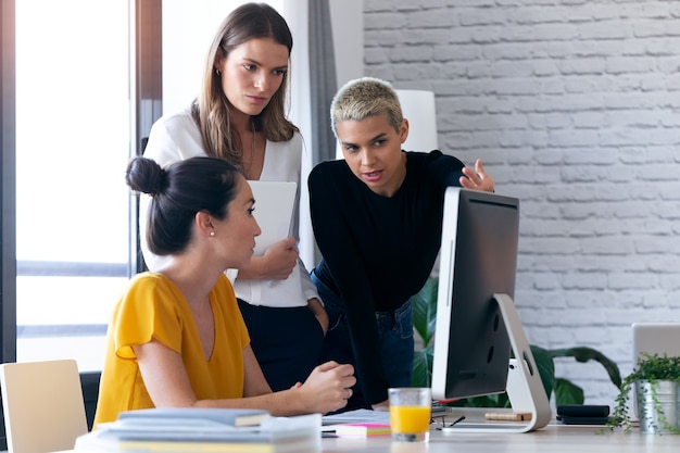 Foto de tres mujeres empresarias modernas hablando y revisando el último trabajo realizado en la computadora en un espacio de trabajo conjunto