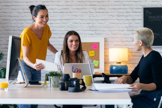 Foto de tres mujeres empresarias modernas hablando y revisando el último trabajo realizado en la tableta digital en un espacio de trabajo conjunto.