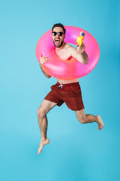 Foto de turista europeo sin camisa hombre vestido con anillo de goma sonriendo mientras juega con pistola de agua juguete aislado