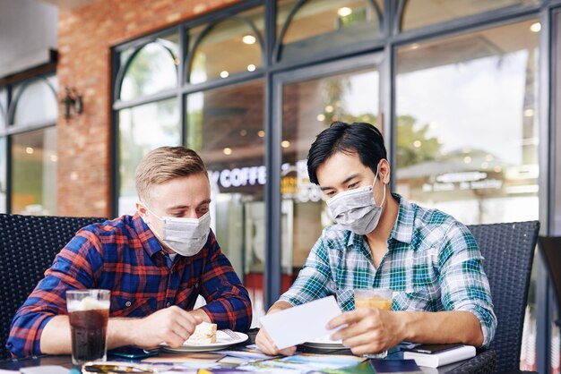 Foto- und Projektmanager in medizinischen Masken treffen sich im Café im Freien, um zu Mittag zu essen und gedruckte Fotos zu besprechen