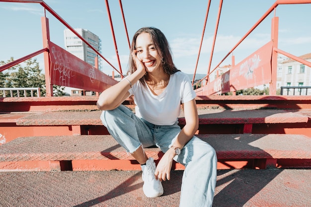 Foto urbana de una joven africana de modelado con cabello largo sentada en un piso rojo mirando seriamente a la cámara. Concepto urbano de la ciudad. Feliz día en la ciudad, camisa blanca, jeans azules. Traje moderno. Copia espacio