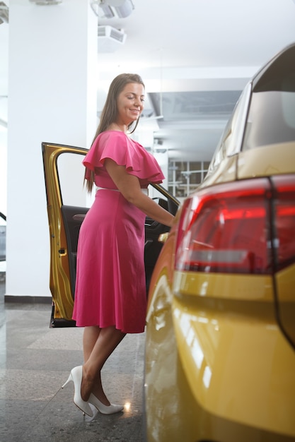 Foto vertical de corpo inteiro de uma mulher feliz examinando um carro à venda na concessionária