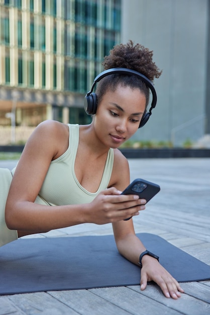 Foto vertical de mulher desportiva descansando no karemat após o treino vestida com roupas esportivas e ouvindo música em hedphones sem fio