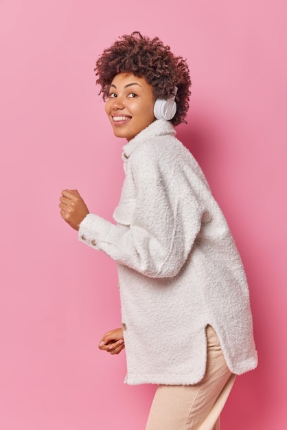 Foto vertical de mulher feliz e despreocupada com cabelo encaracolado dançando e se movendo ao ritmo da música usando jaqueta de pele e calças usando fones de ouvido sem fio isolados sobre a parede rosa