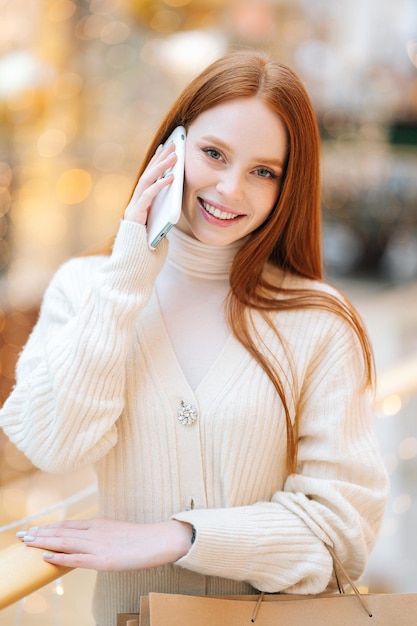 Foto vertical de mulher jovem e atraente confiante falando em um smartphone segurando sacolas de papel