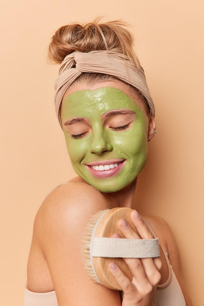 Foto foto vertical de mulher jovem satisfeita aplica máscara facial verde para cuidados com a pele, massagens, corpo com escova usa bandana isolada sobre fundo bege. tempo de beleza e conceito de procedimentos anti-envelhecimento