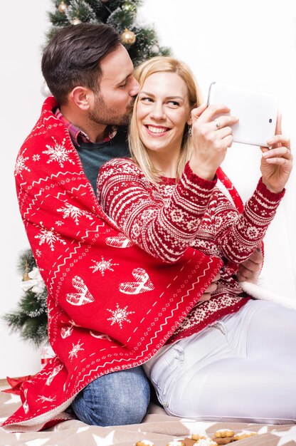 Foto vertical de um casal feliz com um cobertor vermelho tirando uma selfie com uma árvore de natal