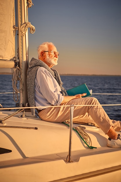 Foto vertical de um homem aposentado relaxado lendo um livro e apreciando o incrível pôr do sol enquanto está sentado no