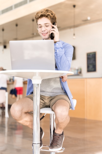 Foto vertical de um jovem com piercings falando em um celular sorrindo sentado em uma mesa com um laptop trabalhando