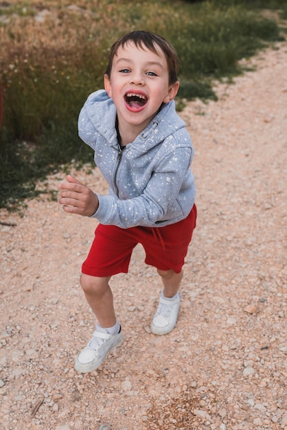 Foto foto vertical de um menino loiro em um moletom cinza e shorts vermelhos correndo alegremente ao ar livre.