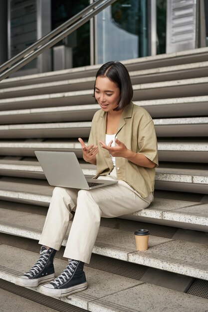 Foto vertical de uma estudante asiática participando de uma reunião on-line conversando em um bate-papo por vídeo em um laptop sentada na escada