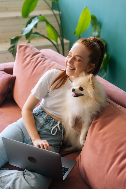 Foto vertical de uma jovem alegre usando laptop sentado em um sofá confortável abraçando um pequeno cão de estimação spitz branco no apartamento