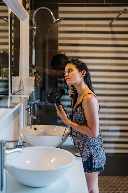 Foto vertical de uma jovem latina secando o cabelo com um secador de cabelo em um banheiro com o marido tomando banho ao fundo