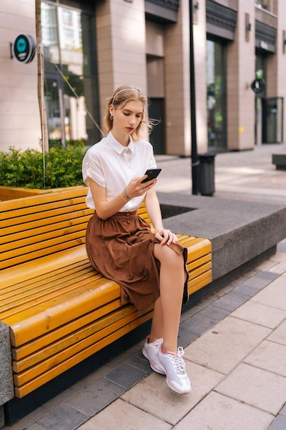 Foto vertical de uma jovem loira séria vestindo roupas casuais sentada no banco amarelo sozinha e verificando as notificações no celular