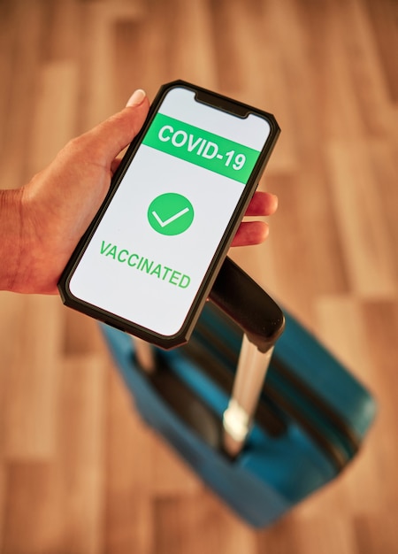 Foto vertical de uma mão de turista mostrando certificado de vacinação de Covid-19 em smartphone com mala para viagem