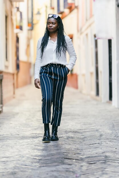 Foto vertical de uma mulher africana com roupas modernas andando distraída por uma rua estreita