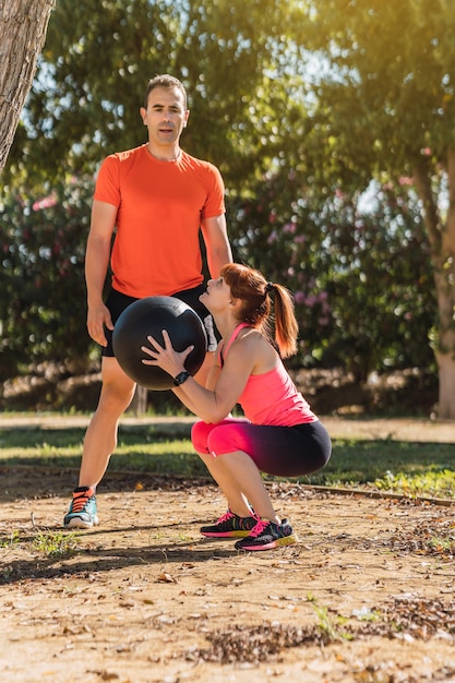 Foto vertical de uma mulher agachada segurando uma bola medicinal com um treinador em um parque