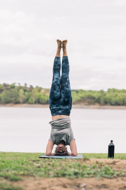 Foto vertical de uma mulher com a cabeça no chão praticando ioga ao lado de um lago em um parque