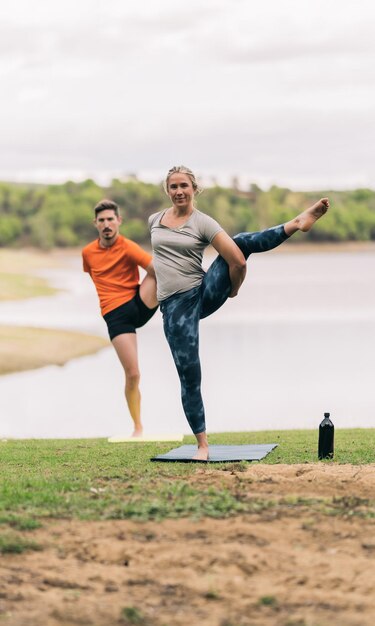 Foto vertical de uma mulher e um homem levantando a perna enquanto fazem ioga ao lado de um lago em um parque