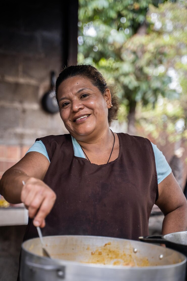 Foto vertical de uma mulher sorridente de avental fritando comida em uma cozinha rural comunitária ao ar livre