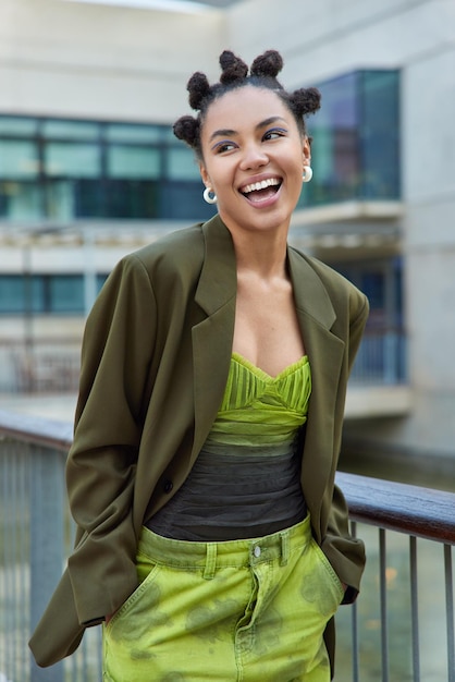 Una foto vertical de una feliz mujer de moda vestida con ropa elegante mantiene las manos en los bolsillos y se ríe felizmente disfruta de poses de tiempo libre al aire libre contra un fondo borroso. Concepto de estilo de vida urbano