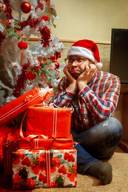 Foto vertical de hipster triste con muchos regalos cerca del árbol de Navidad. Año nuevo.