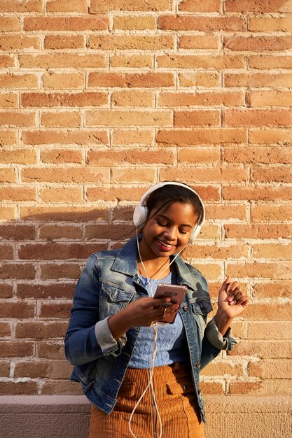 Foto vertical de joven afroamericana con auriculares escuchando música, cantando y bailando