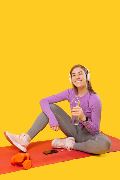 Foto vertical de una joven deportista que usa audífonos y sostiene una botella de agua y se sienta en una estera Foto de estudio sobre fondo amarillo