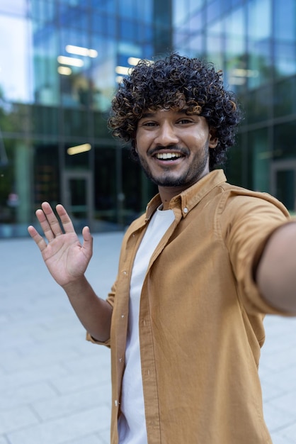 Foto vertical joven estudiante hispano sonriente hablando por videollamada a la cámara de pie afuera