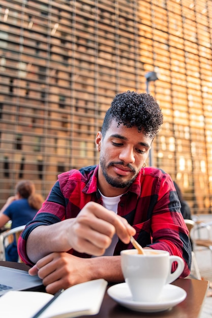 Foto vertical de un joven latino revolviendo su café con una cuchara mientras trabajaba en la mesa de la terraza de una cafetería, concepto de estilo de vida urbano