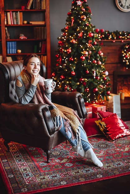 Foto vertical de una linda mujer sonriente sentada en un sillón con una taza en las manos mirando la cámara