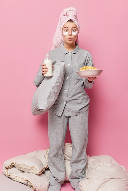 Una foto vertical de una mujer joven y bonita mantiene los labios doblados vestidos con un pijama gris, sostiene una botella de almohada de leche y cereales, se despierta por la mañana y desayuna aislado sobre un fondo rosa.