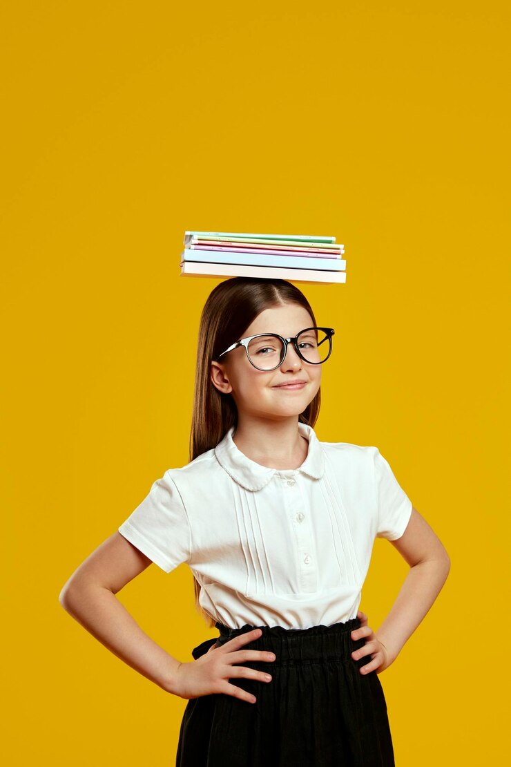Foto vertical de una niña juguetona de pie con confianza con las manos en las caderas equilibrando libros en la cabeza