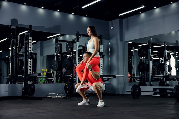 Una foto de la vibración positiva de una pareja de fitness. El hombre está en cuclillas y la niña sentada sobre sus hombros en un gimnasio oscuro. Objetivo de pareja, apoyo físico